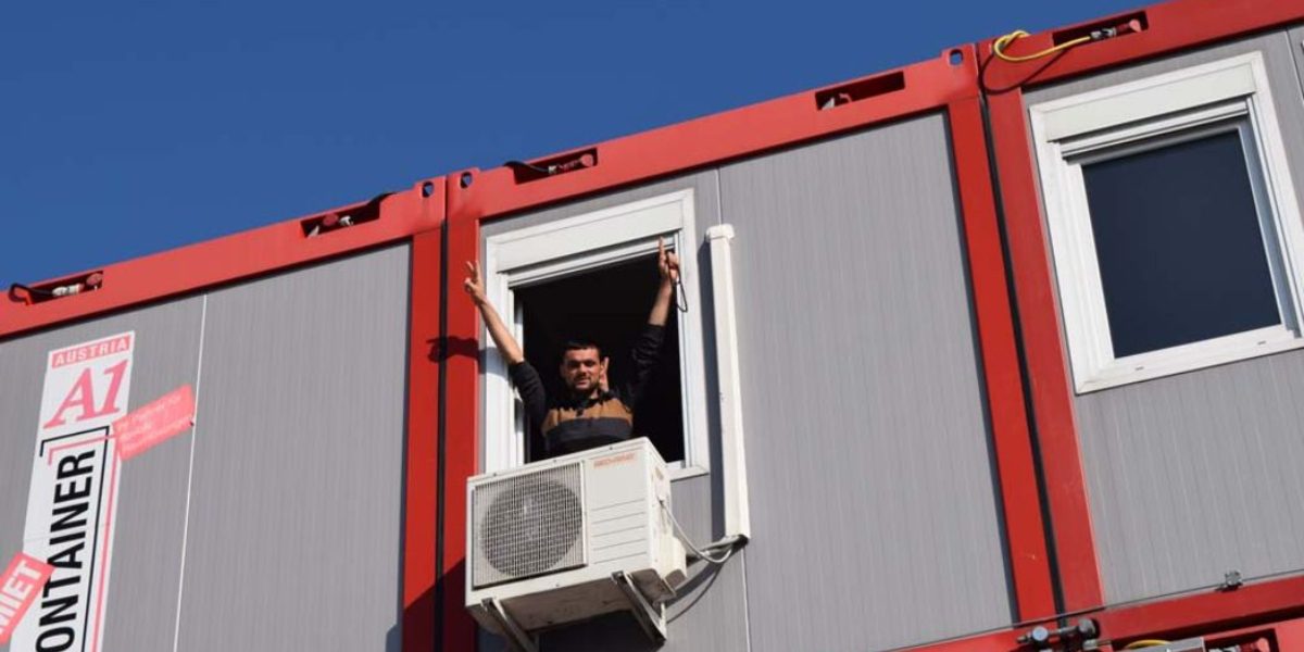 Un joven hace el signo de la paz desde un contenedor convertido en casa. Muchos refugiados están alojados en este tipo de viviendas provisionales en Europa. (Servicio Jesuita a Refugiados)